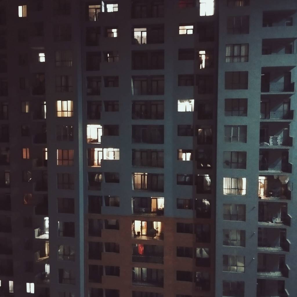 Ein großes Hochhaus in der Nacht, beleuchtet von verschiedenen Fenstern, die ein Gefühl von Leben und Aufeinanderprall von Privatsphäre und Beobachtung vermitteln.