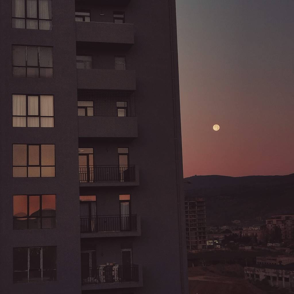 Blick auf ein Hochhaus mit mehreren Balkonen und Fenstern, während der Vollmond am Horizont aufgeht und der Himmel in warmen Farben leuchtet.