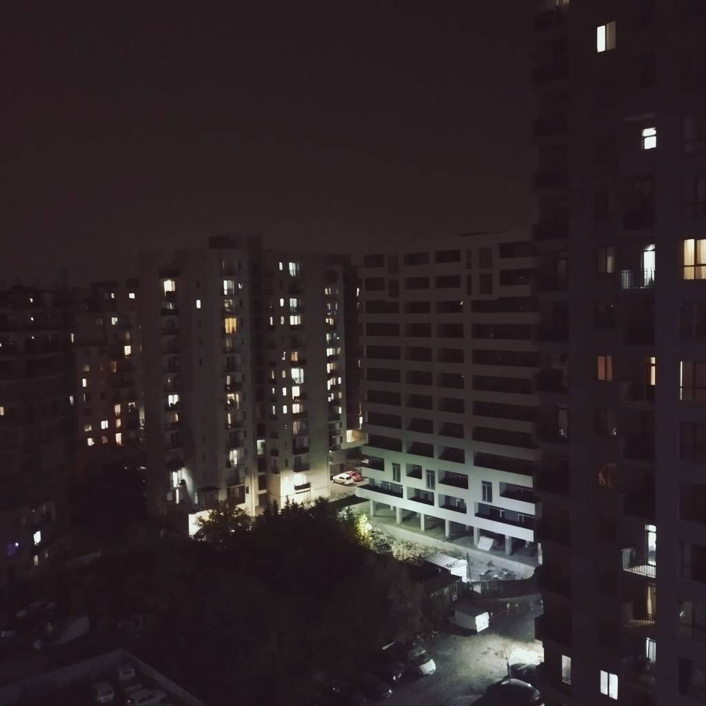 Nachtaufnahme eines Wohngebiets mit mehreren Hochhäusern, deren Fenster teilweise beleuchtet sind, und einem Blick auf den darunterliegenden Parkplatz.